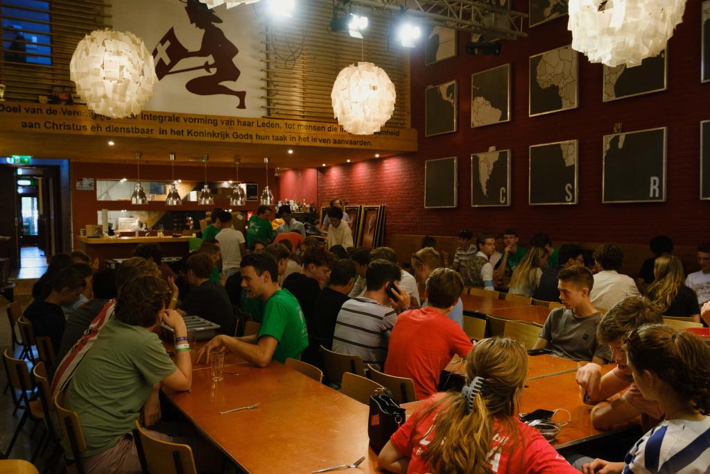 Grote , sfeervolle ruimte van een studentensociëteit gevuld met biertafels waaraan studenten zitten, in de achtergrond is de bar.