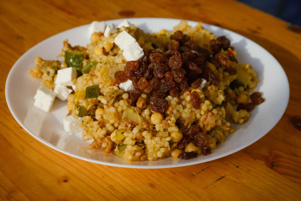 Wit bord met parelcouscous met rozijnen, groente en blokjes feta op een houten biertafel.