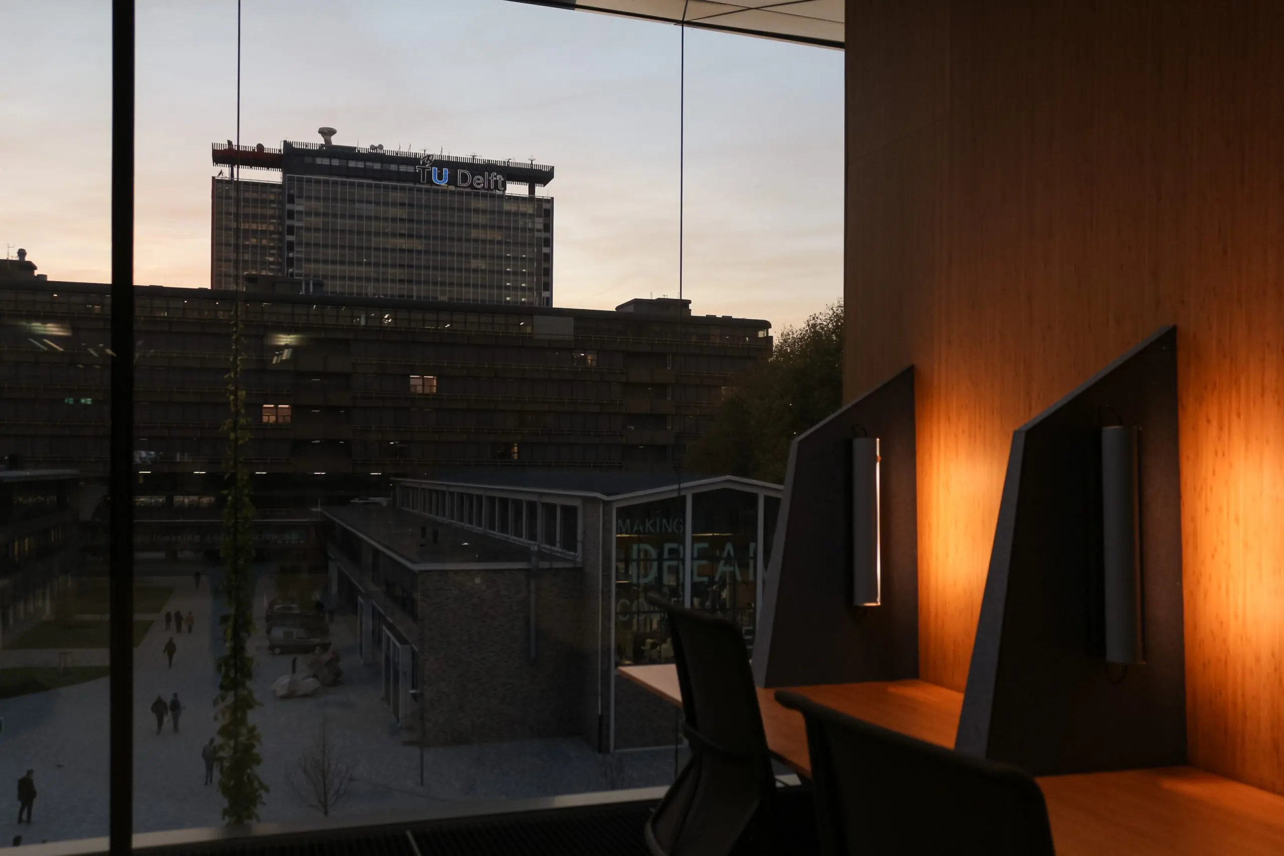 Rechts twee studieplekken met verlichting. de rest van de foto wordt in beslag genomen door een glazen pui, waarachter gebouwen van de TU Delft zijn te zien. Eentje draagt het logo van de TU Delft