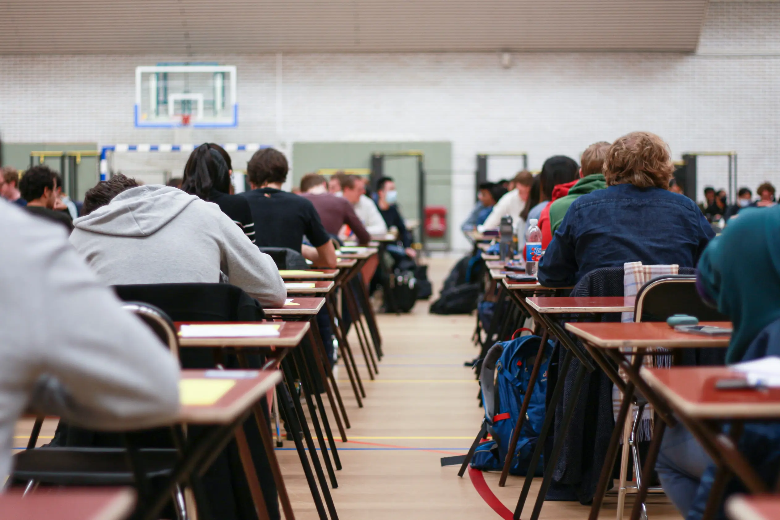 TU, steun dubbelstudenten Twee rijen mensen, van achteren gezien, zitten gebogen over kleine bureaus in een sporthal