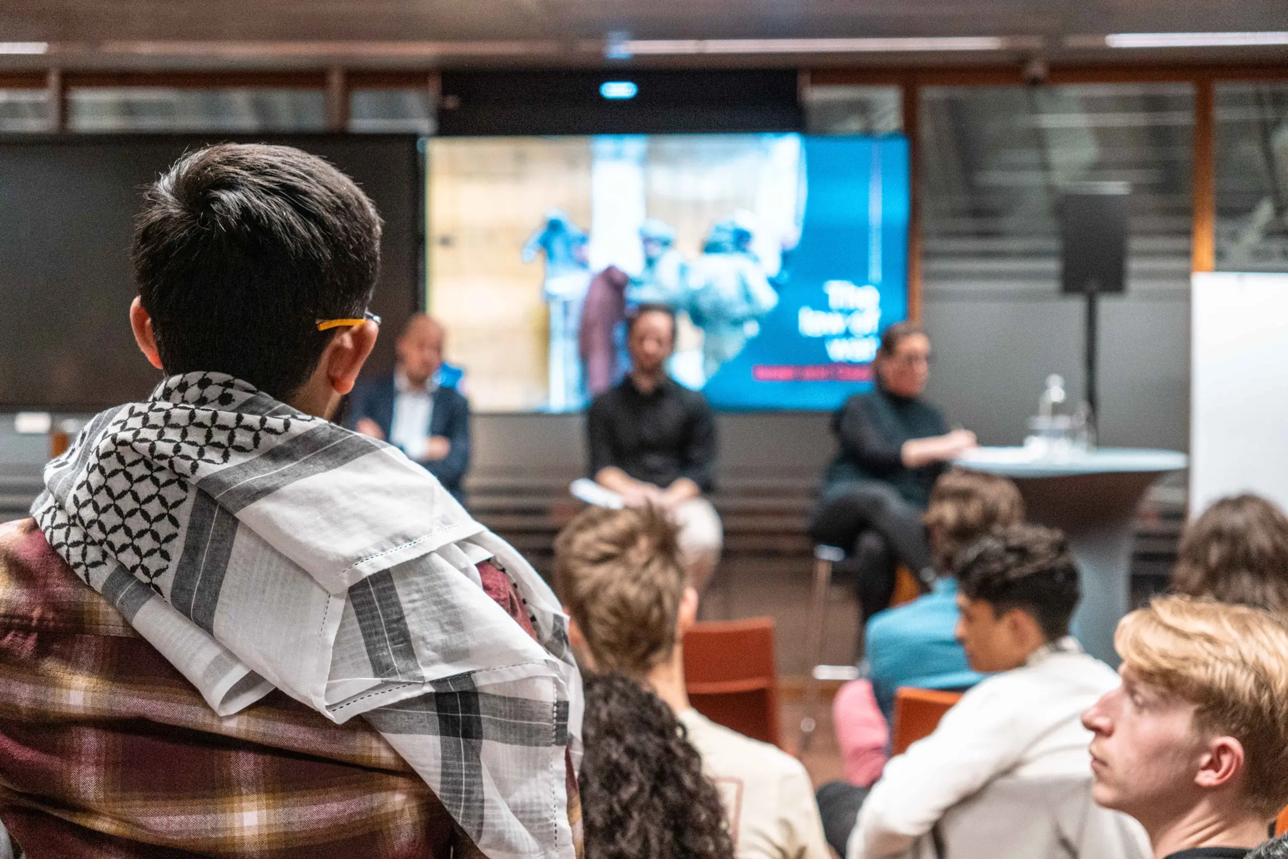 Een publiek, op de rug gezien, kijkt naar drie mensen op krukken. Op de voorgrond een jongeman van achteren gezien, met een kufiya om
