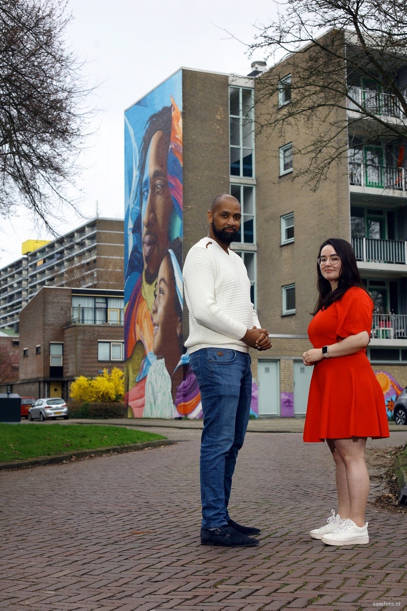 Een foto van een beschilderd flatgebouw met op de voorgrond een man en een vrouw die de muurschildering mede-bedachten.