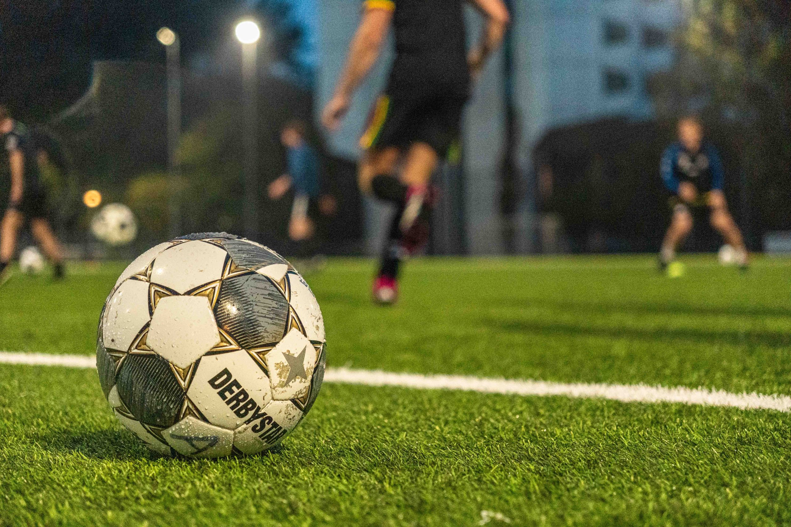 Een voetbal op een door lampen verlicht veld. Vaag in de achtergrond rennen vier voetballers