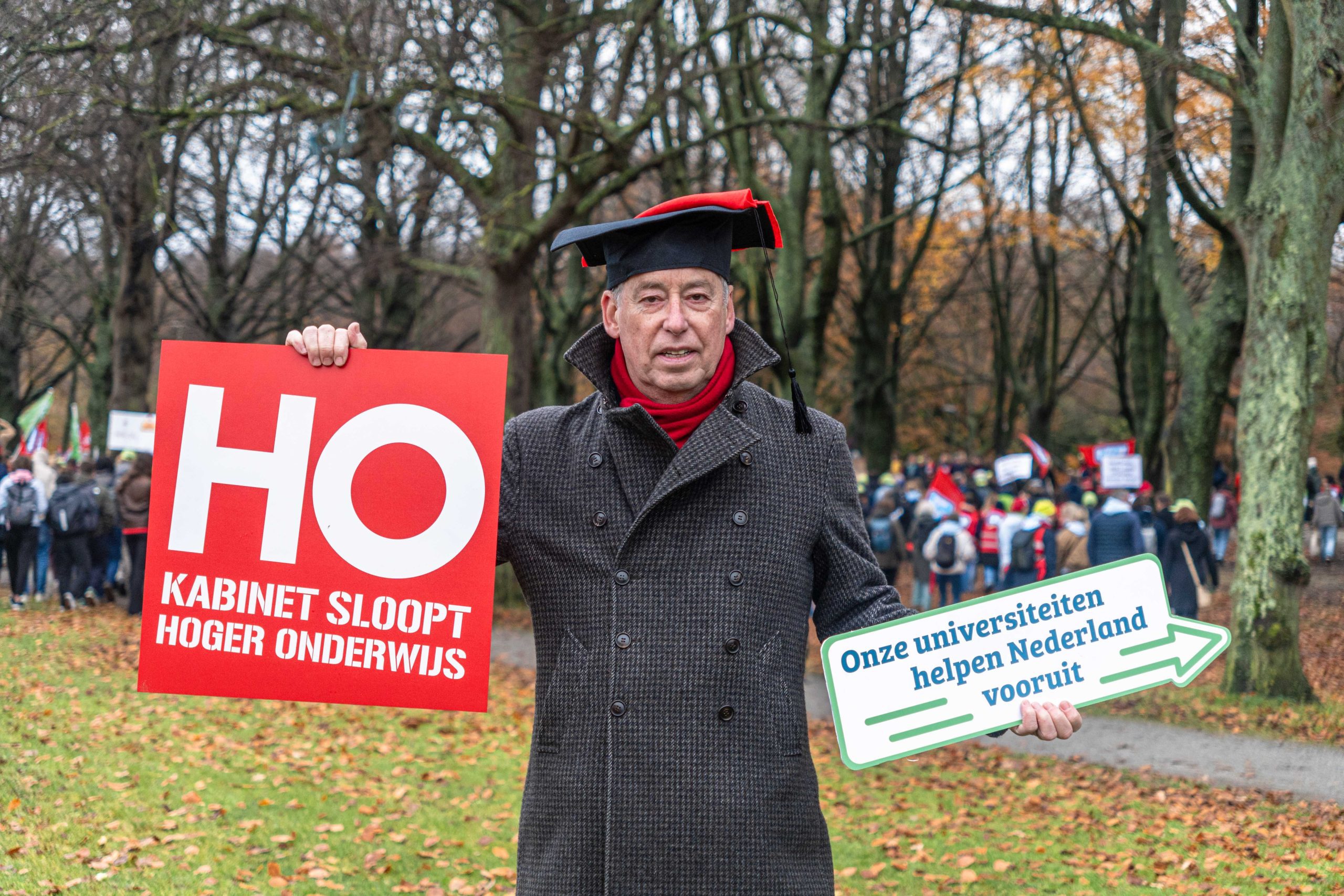 Rector magnificus Tim van der Hagen poseert voor de foto. Op zijn hoofd een doctoraalhoed met een rood vierkant, een teken van WOinActie. Hij heeft twee borden vast. Op de een staat: HO, Kabinet sloopt hoger onderwijs. Op de ander staat: Onze universiteiten helpen Nederland vooruit