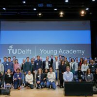 Een groep mensen poseert voor de foto. Ze staan op een podium. Achter hen een beeldscherm met de tekst Delft Young Academy
