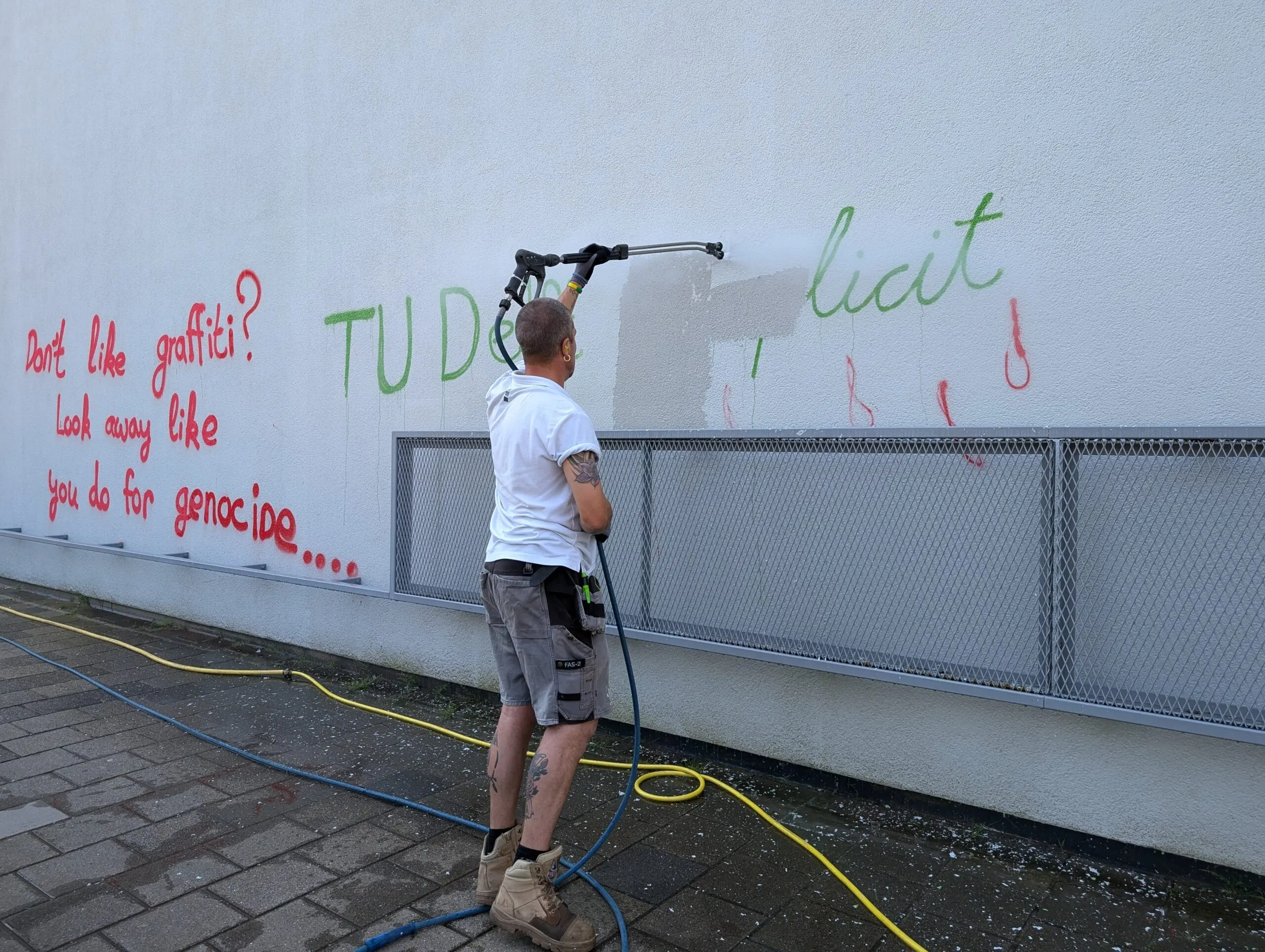 Een man verwijdert met hoge drukspuit rode en groene graffiti van een grijze muur.