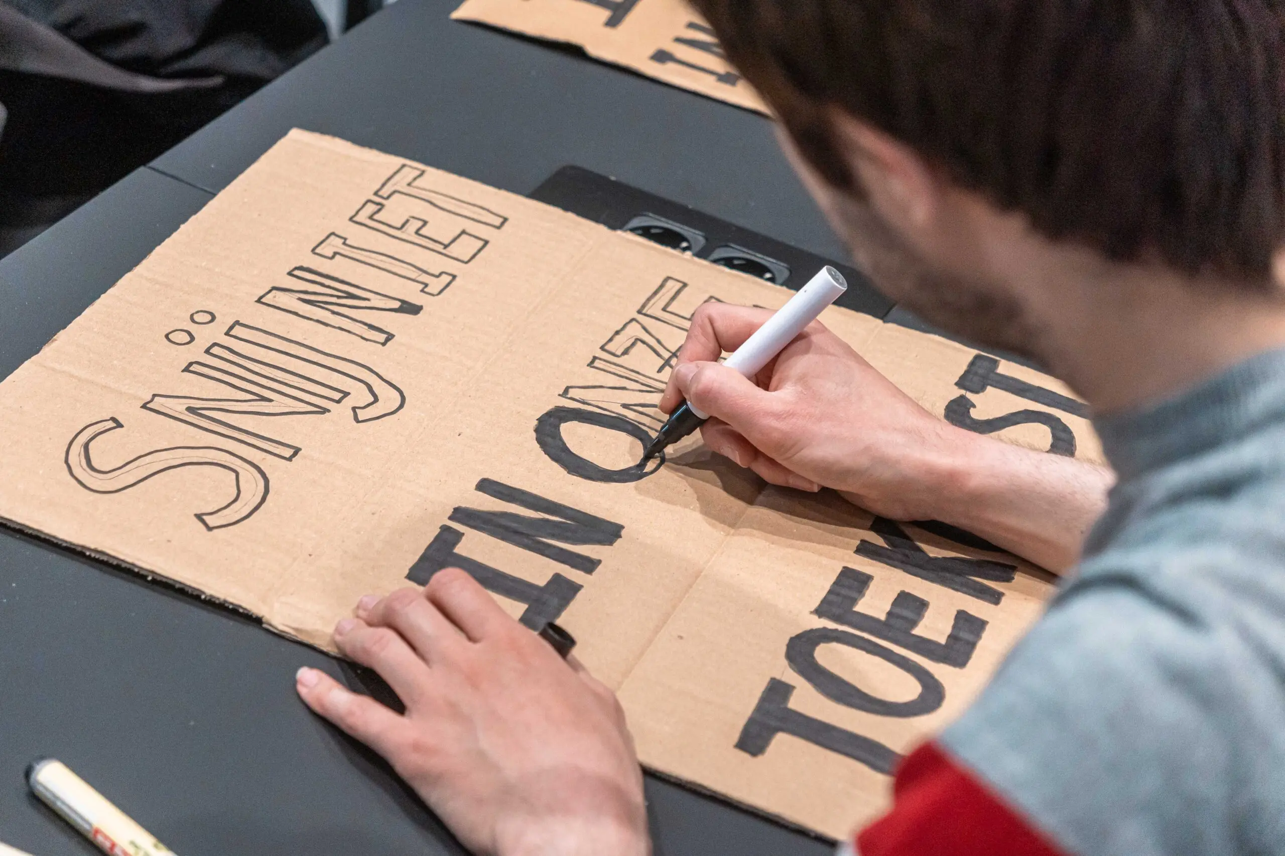 Close up van iemand die niet met het gezicht te zien is die een protestbord schrijft met de tekst Snij niet in onze toekomst