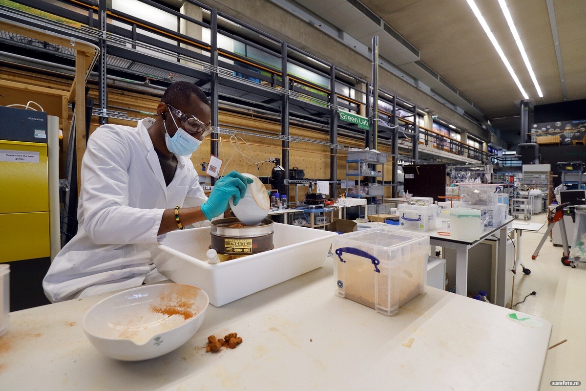 Iemand in een witte labjas, een mondkapje, een beschermende bril en handschoenen is aan het werk in het lab