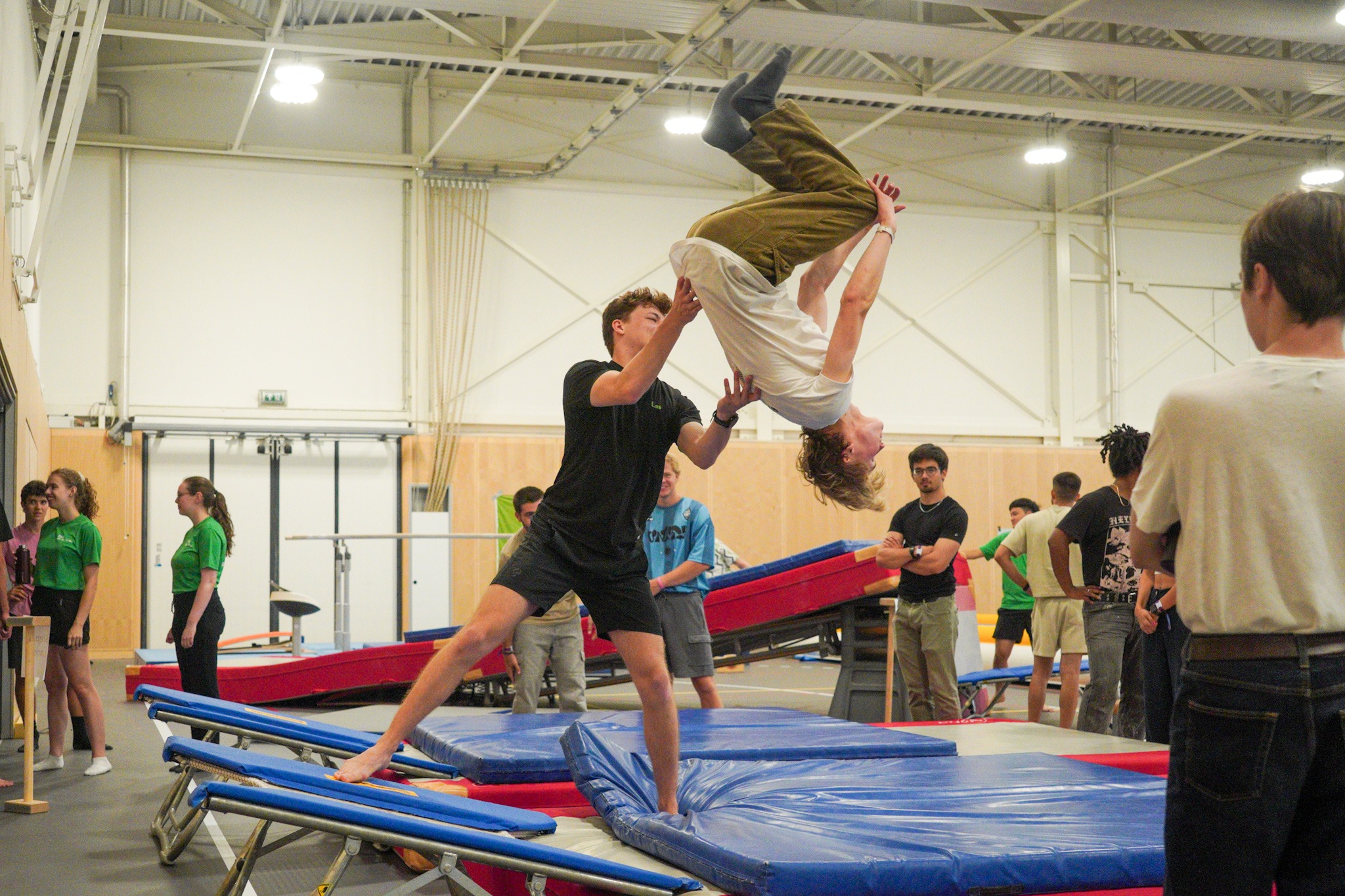 Een jongen maakt een achterwaartse salto vanaf een trampoline terwijl een student die ernaast staat hem helpt draaien.