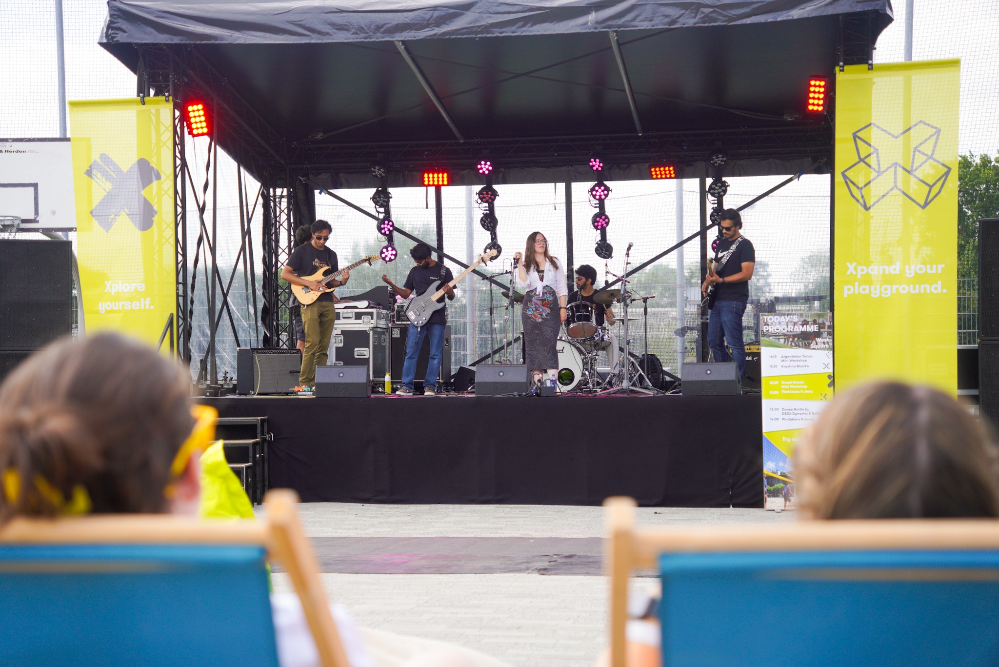 Een band staat op een tijdelijk podium op het terrein van X. Onscherp in de voorgrond zitten twee mensen in strandstoelen te luisteren.
