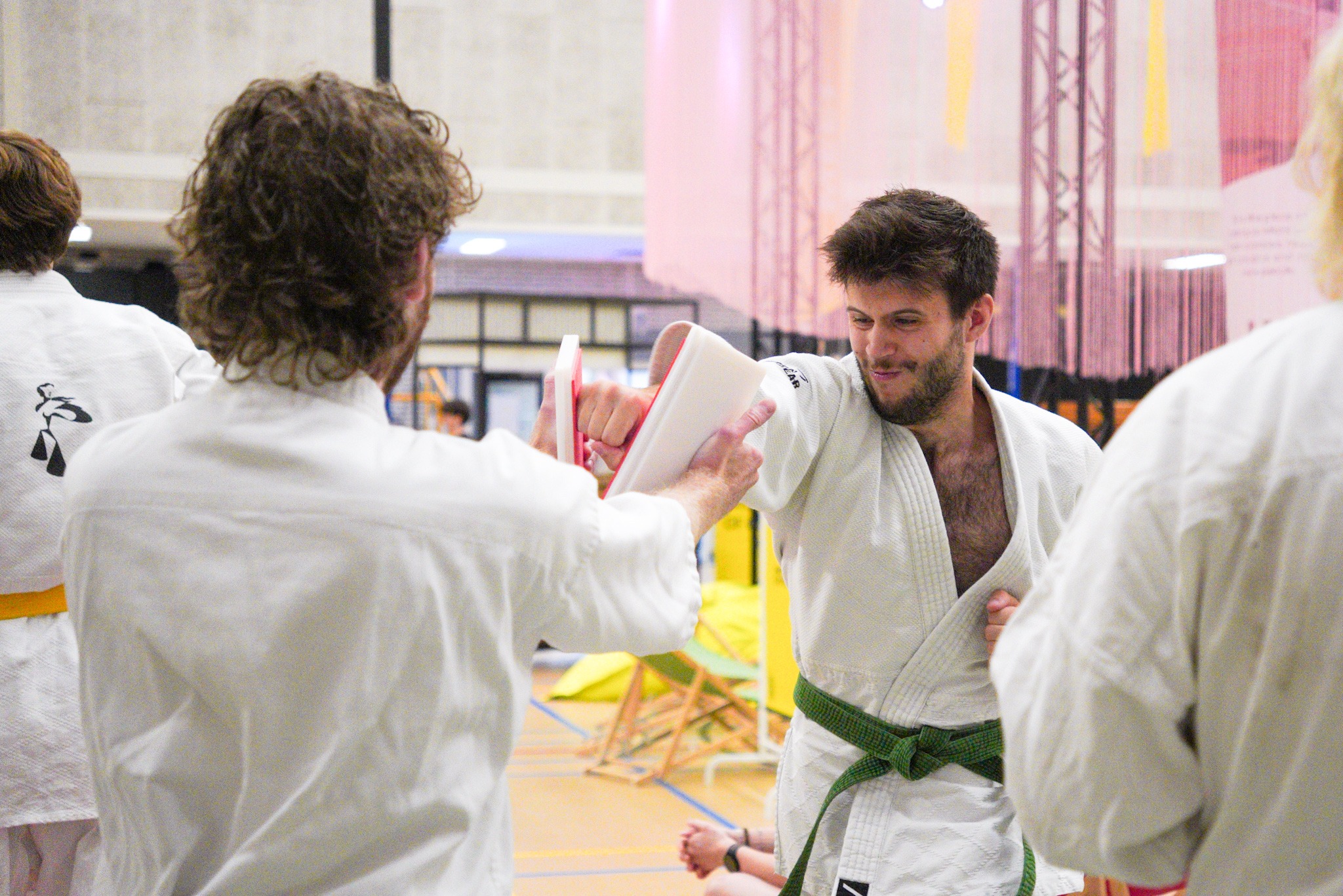 Iemand in judokleding staat met een gestrekte arm met zijn vuist tussen twee helften van een plankje die door iemand anders worden vastgehouden.