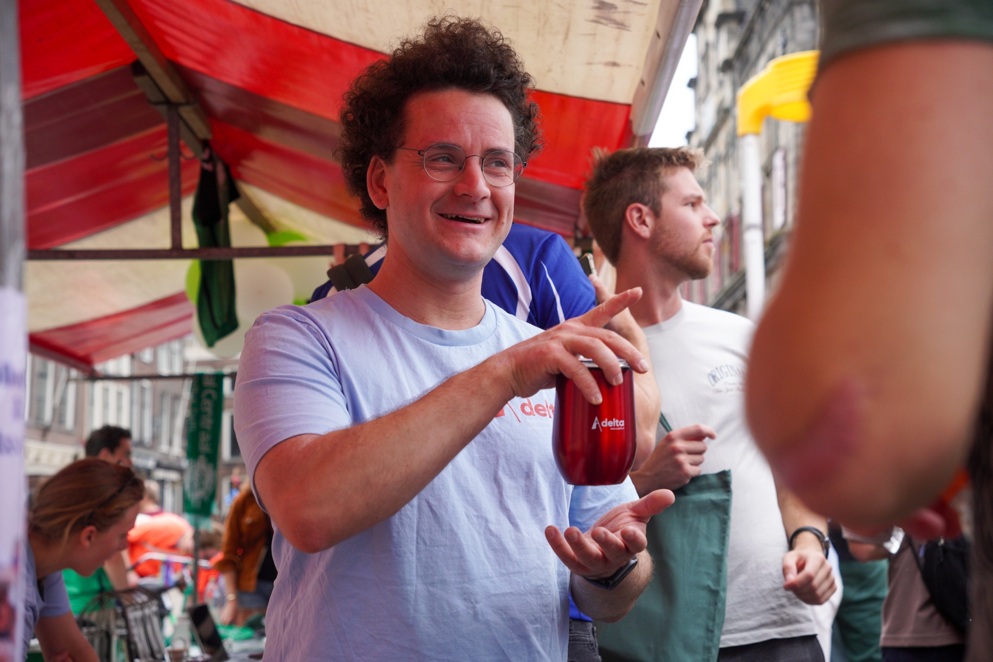 Delta-redacteur Emiel Beinema draagt een t-shirt van Delta en heeft een mok van Delta in zijn handen terwijl hij bij een marktkraam in gesprek is met iemand buiten beeld.