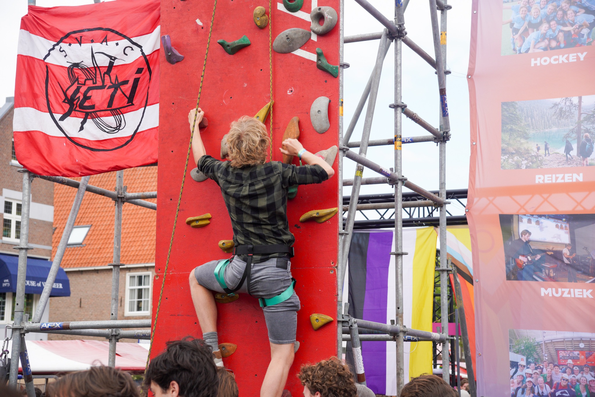 Tegen een steiger hangen een klimwand en een vlag van S.V.A.C. Yeti. Een man klimt, gezekerd aan een touw, omhoog.
