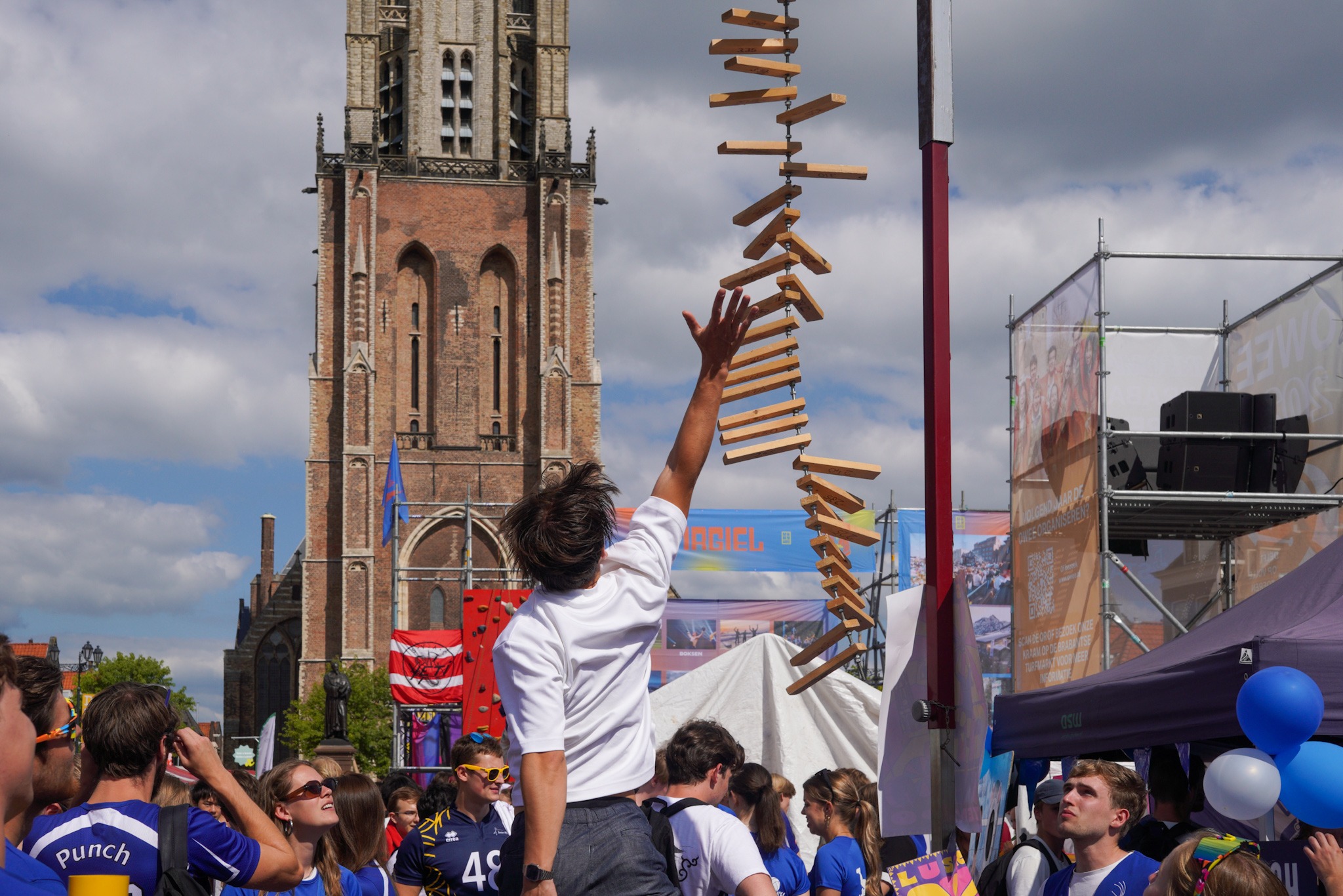 Een man in een wit t-shirt springt om zo hoog mogelijk een houten latje aan te tikken. Studenten in t-shirts van volleybalvereniging Punch staan eromheen en kijken toe.