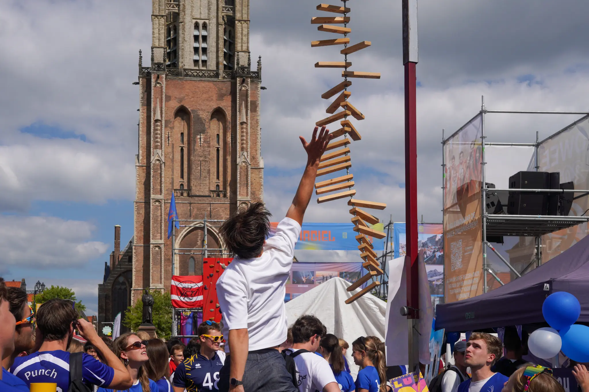 Een man in een wit t-shirt springt om zo hoog mogelijk een houten latje aan te tikken. Studenten in t-shirts van volleybalvereniging Punch staan eromheen en kijken toe.