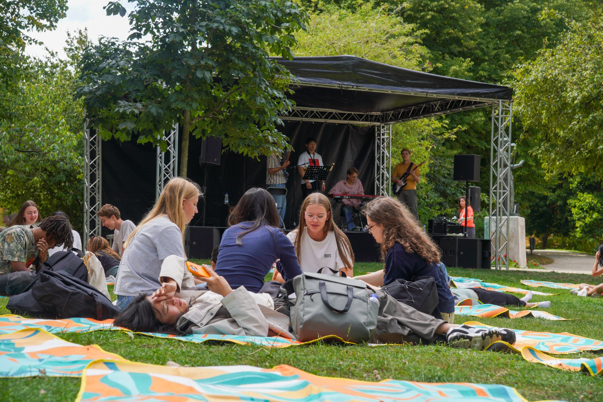 Een groepje deelnemers aan de OWee zit in het gras terwijl achter hen op een overdekt podium een bandje speelt.