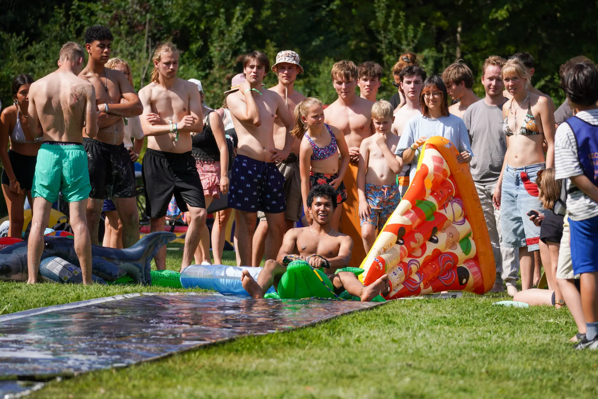 Een OWee-deelnemer zit lachend op een opblaaskrokodil aan het begin van een zeepbaan. Achter hem staat een groep toeschouwers, sommigen in zwemkleding. Een persoon houdt een opblaas-pizzapunt vast.