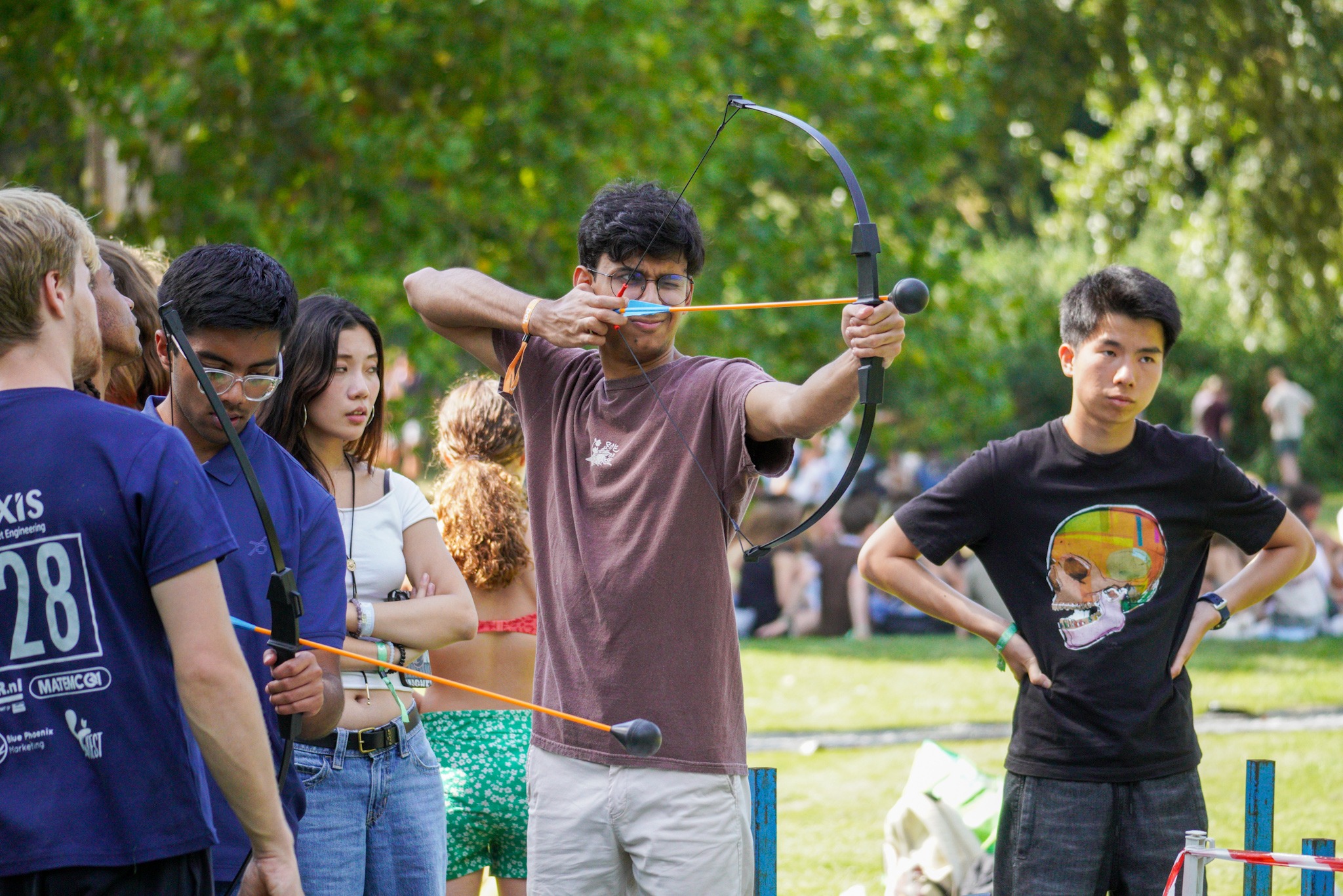 Een deelnemer aan de OWee staat met gespannen boog, klaar om te schieten. Andere deelnemers staan om hem heen en kijken toe.