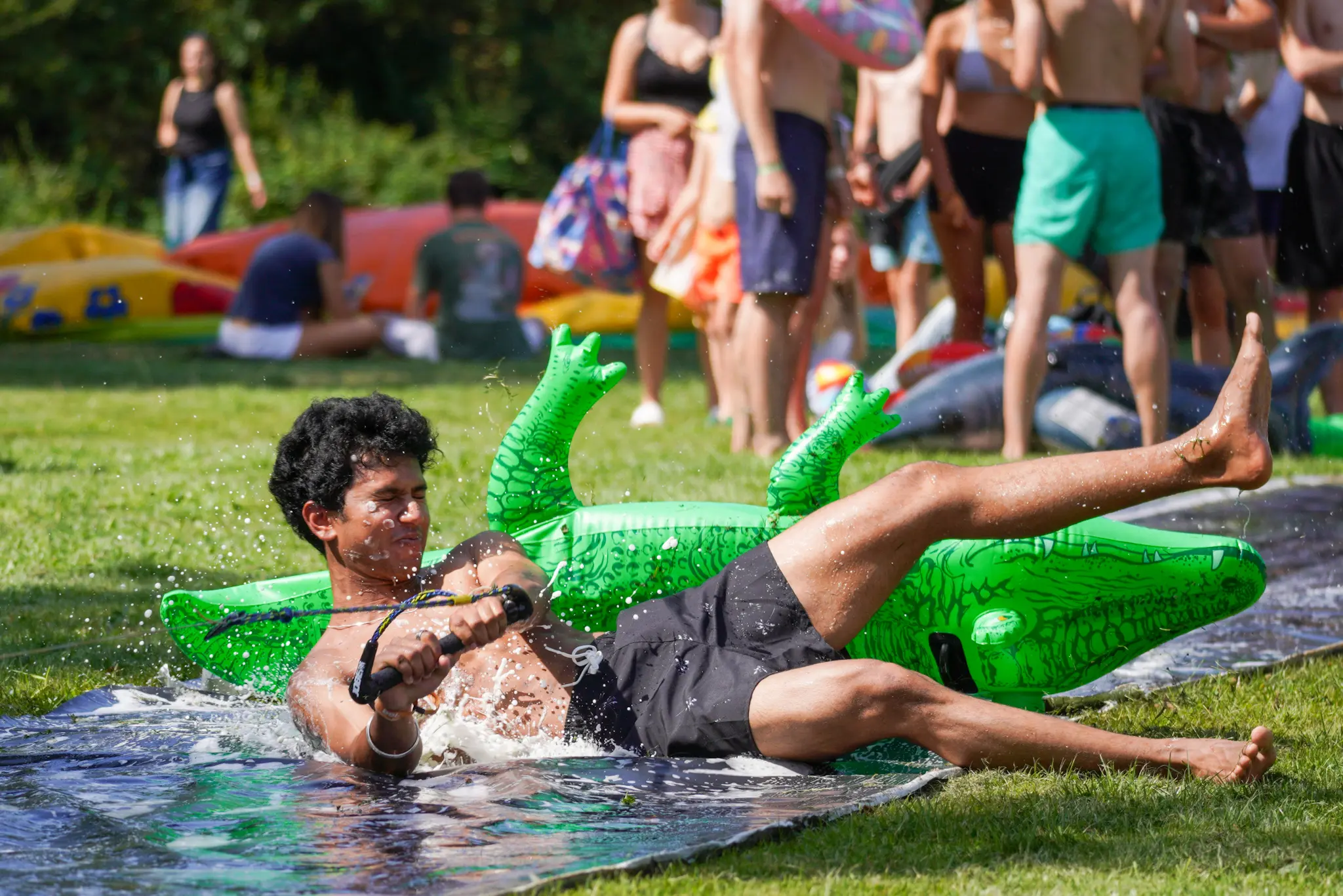 Een deelnemer aan de OWee houdt zich vast aan een touw waarmee hij over een zeepbaan wordt getrokken. Achter hem ligt een opblaaskrokodil.