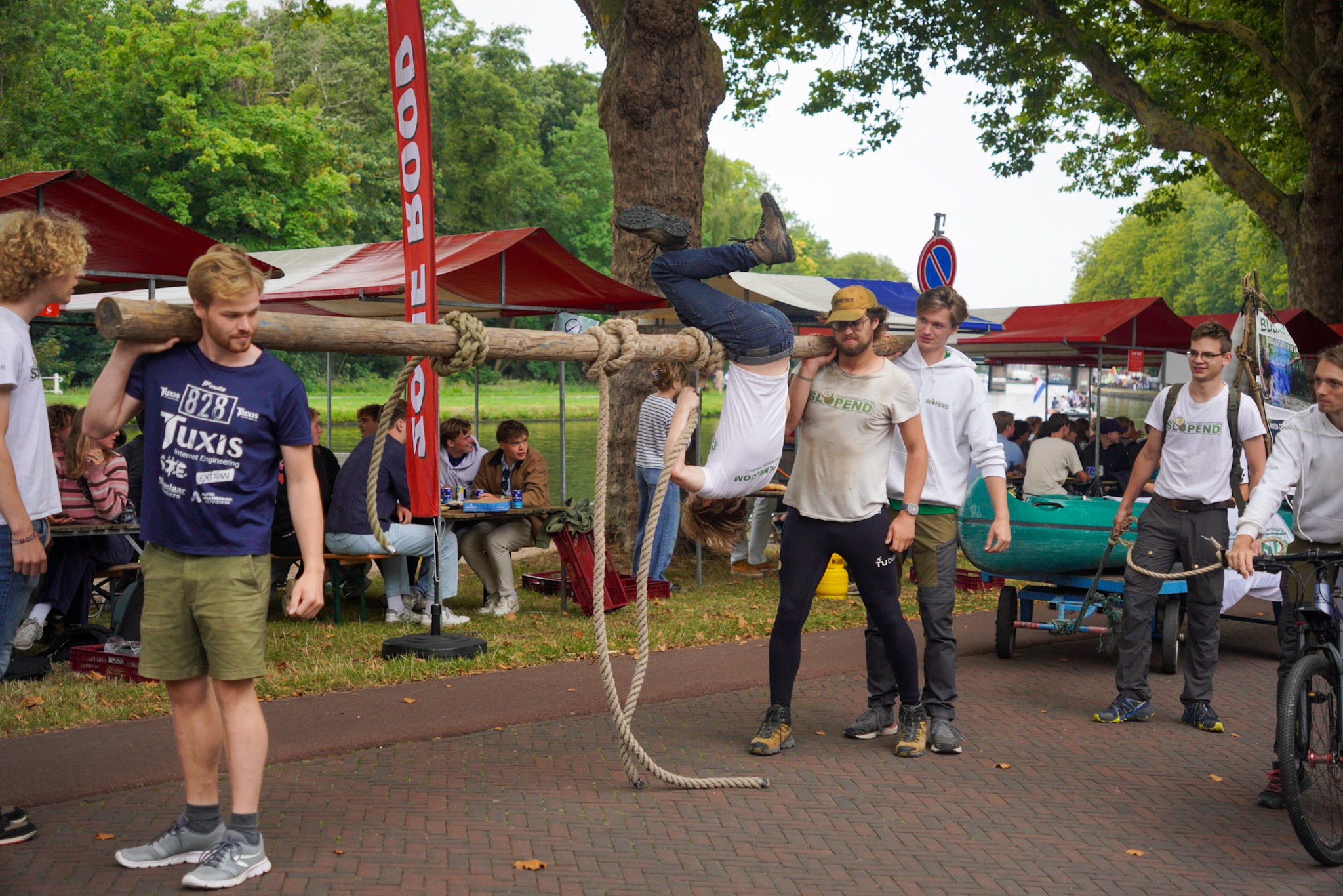 Twee mannen dragen een boomstam, een derde man hangt daar op de kop aan.