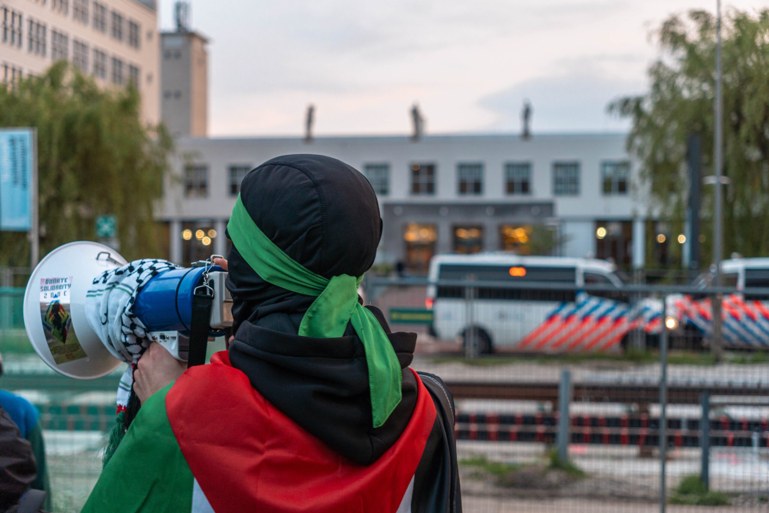 Actiegroep opent eigen meldpunt voor incidenten rondom Palestina op TU Delft Een demonstrant met een Palestijnse vlag om de schouders en een kap over het hoofd spreekt in een versterker. Op de achtergrond staan twee politiebussen