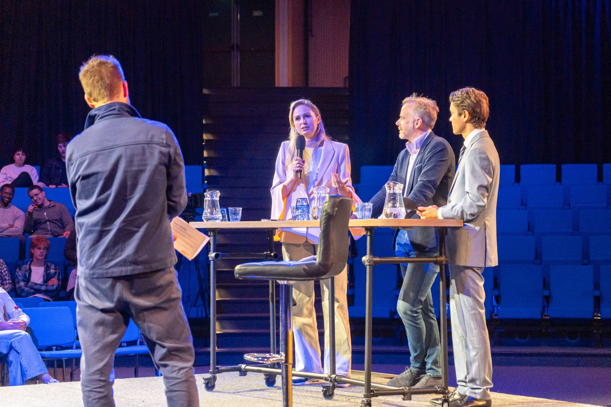 Queeny Rajkowski (VVD), Joost van der Sluis (SP) en Dion Huidekooper (D66) staan achter een tafel te debatteren. Rajkowski houdt een microfoon vast en praat tegen moderator Merijn Doggen, die met zijn rug naar de fotograaf staat.
