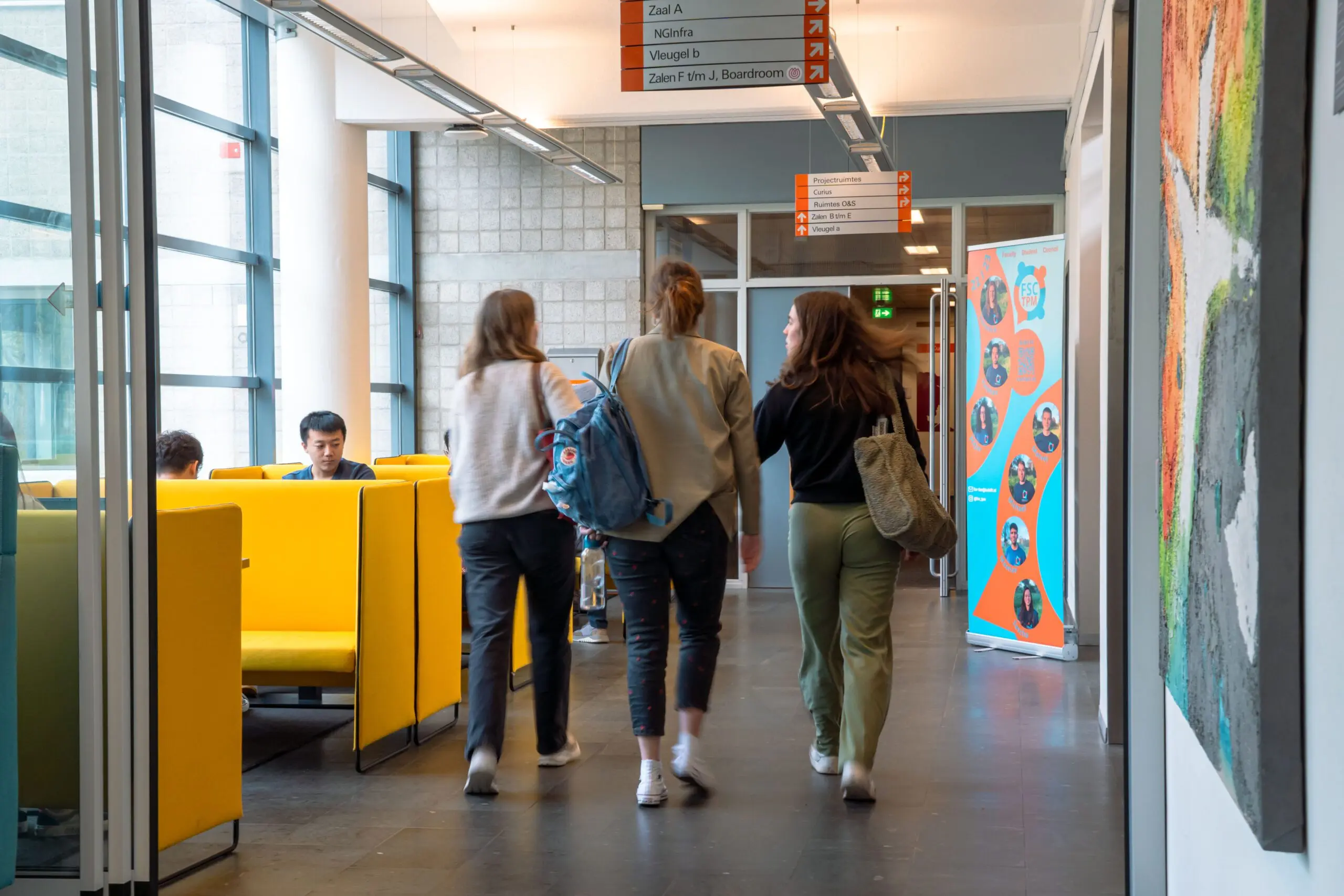 How should we behave, TU Delft asks Drie mensen lopen door een gang. Je ziet ze op hun rug