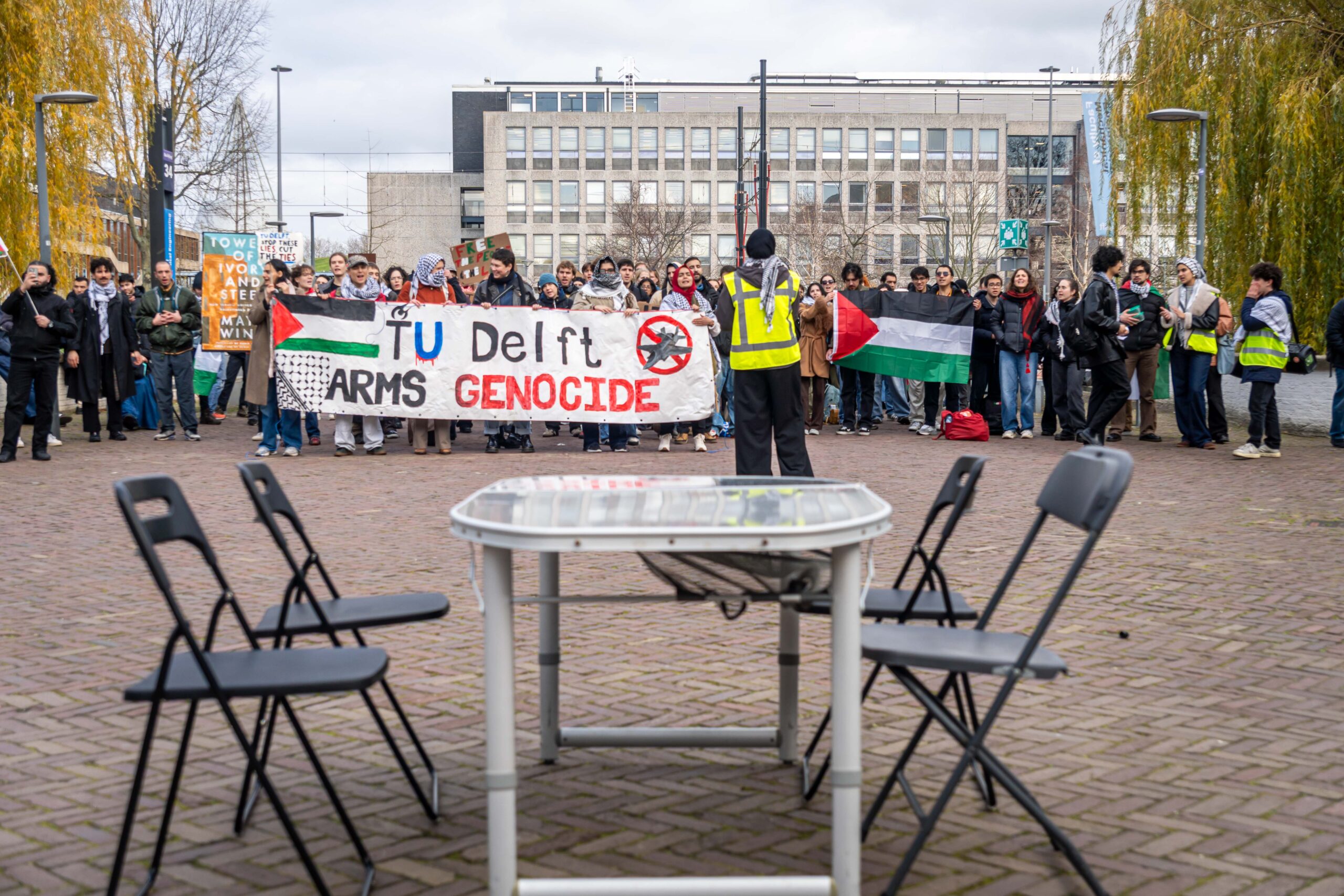 Op de voorgrond een tafeltje met 4 klapstoelen, op de achtergrond een groep demonstranten. Ze dragen een spandoek met de tekst 'TU Delft arms genocide'