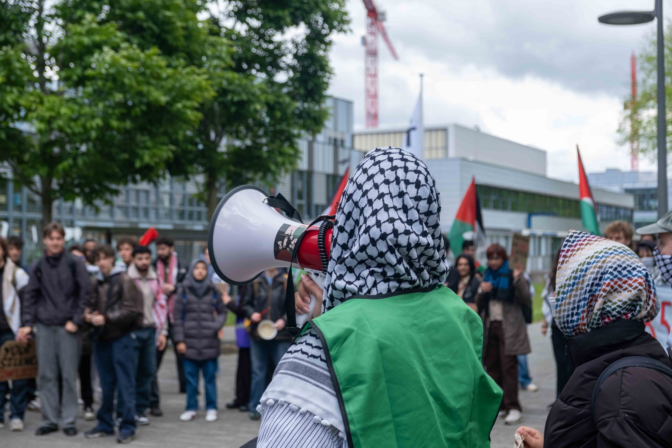 demonstrant met keffiyeh spreekt door een megafoon. OP de achtergrond staan mensen te luisteren