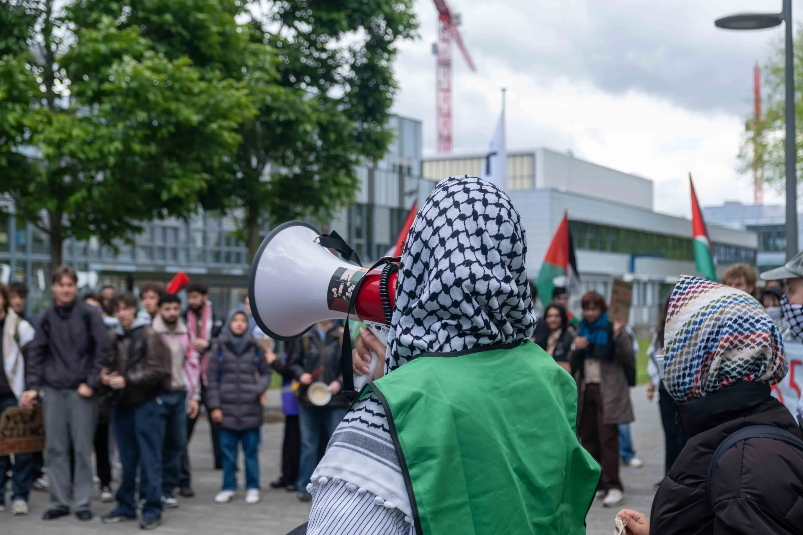 demonstrant met keffiyeh spreekt door een megafoon. OP de achtergrond staan mensen te luisteren