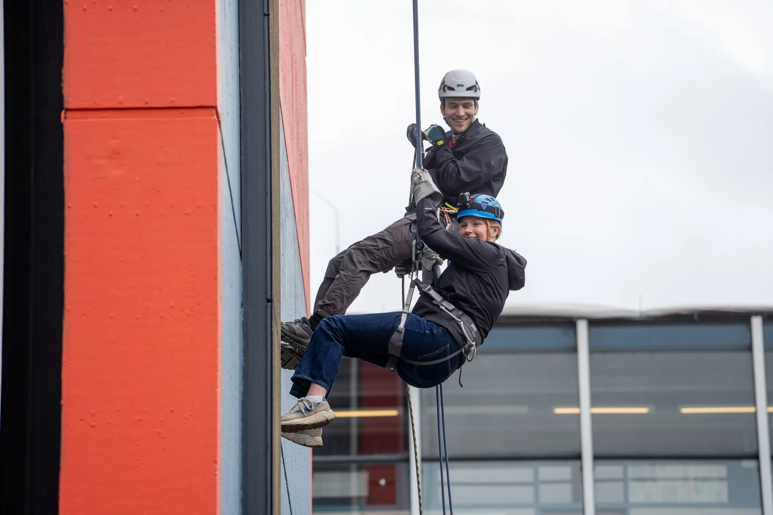 twee abseilers hangen aan touwen langs de EWI hoogbouw en lachen naar de camera